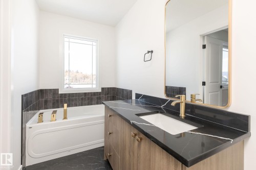 Built-in soaking tub with gold-finish fixtures, wood-finish vanity with black countertop and integrated sink, and an accent wall with dark subway tile - 24 Clichy Close, St. Albert, AB - Indoor Photo Showing Bathroom