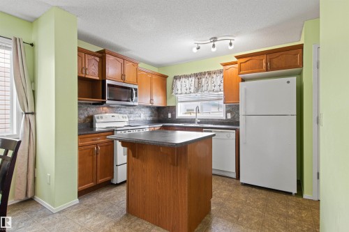 Kitchen featuring wood-finish cabinetry, a central island with a dark countertop, tile flooring, and white appliances including a refrigerator, range, and dishwasher - 9118 Scott Crescent Nw, Edmonton, AB - Indoor Photo Showing Kitchen