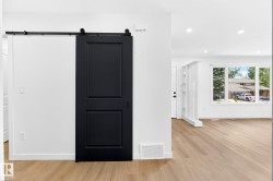 Black barn door with exposed hardware, wood-finish flooring, white walls, recessed ceiling lighting, and a large window - 