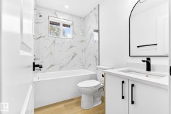 White vanity with integrated sink and matte black faucet, framed arched mirror, white subway tile with gray veining, white bathtub with matte black fixtures, and wood-finish flooring - 