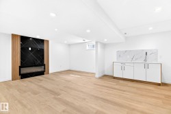 Wood-finish flooring throughout, recessed ceiling lighting, a built-in linear fireplace with a black accent wall and wood paneling, and a wet bar with white cabinetry and a patterned backsplash - 