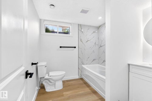 Bathroom featuring a bathtub with a tiled surround, a toilet, a window, wood-finish flooring, and a vanity with a circular mirror - 5 Peace River Drive, Devon, AB - Indoor Photo Showing Bathroom