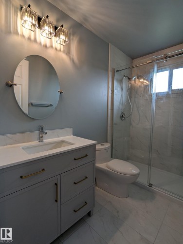Vanity with a white countertop and gold-toned hardware, oval mirror, crystal-style vanity lighting, glass-enclosed shower, and large format floor tiling - 1907 145A Avenue Nw, Edmonton, AB - Indoor Photo Showing Bathroom