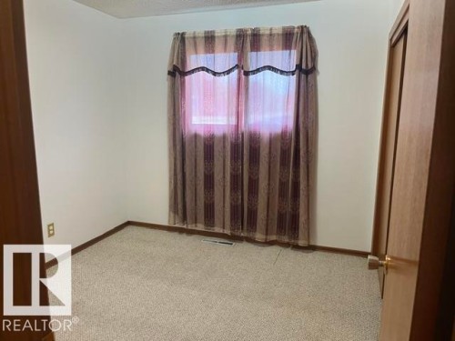 Carpeted room featuring a window with patterned drapes, white walls, and wood-finish trim - 5213 50 Avenue, Vegreville, AB - Indoor Photo Showing Other Room
