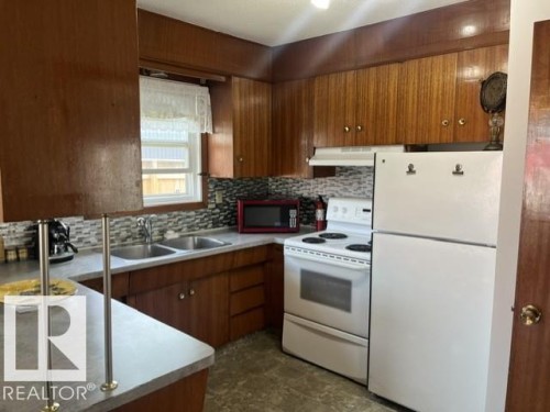 Kitchen featuring wood-finish cabinetry, a double basin stainless steel sink, and a multi-tone tile backsplash - 5213 50 Avenue, Vegreville, AB - Indoor Photo Showing Kitchen With Double Sink