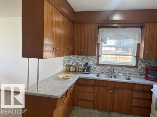 Kitchen featuring wood-finish cabinetry, a dual basin stainless steel sink, and a multi-toned mosaic tile backsplash - 5213 50 Avenue, Vegreville, AB - Indoor Photo Showing Kitchen With Double Sink
