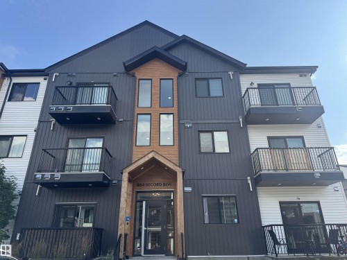 Modern building exterior featuring dark panel siding, multiple balconies with dark railings, large windows, and a prominent wood-clad entryway - #301 864 Secord Bv Nw, Edmonton, AB - Outdoor With Balcony