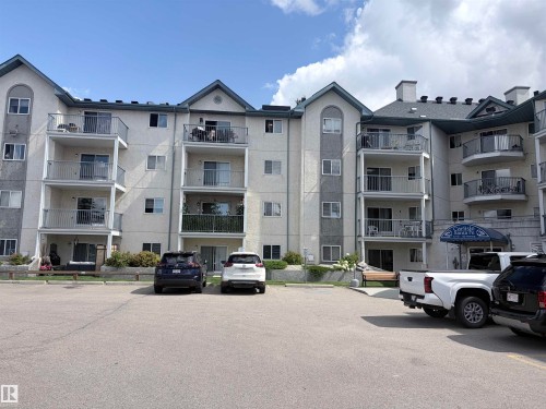 Multi-story residential building with stucco and stone veneer exterior, multiple private balconies with metal railings, a central entrance with an awning, and a paved parking area - #208 6710 158 Av Nw, Edmonton, AB - Outdoor With Balcony With Facade