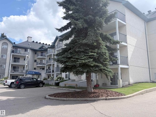 Multi-story building featuring stucco exterior, multiple balconies with metal railings, a paved parking area, and mature landscaping - #208 6710 158 Av Nw, Edmonton, AB - Outdoor With Balcony