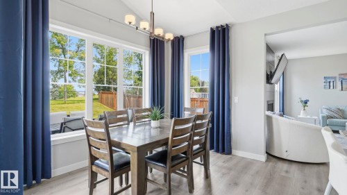 Bright dining area with expansive multi-pane windows overlooking manicured grounds, navy grommet-top curtains, wood-finish flooring, pendant lighting fixture, and direct sightlines to an adjacent kitchen with white cabinetry and stainless steel appliances - 63 Rosemount Boulevard, Beaumont, AB - Indoor Photo Showing Dining Room