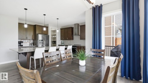 Open-concept dining and kitchen area featuring dark wood-finish flooring, stainless steel appliances, pendant lighting, navy blue drapery panels, and abundant natural light from multiple windows and glass doors - 63 Rosemount Boulevard, Beaumont, AB - Indoor