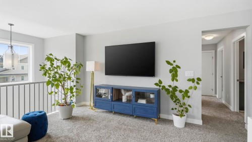 Modern living room featuring a wall-mounted flat-screen television, blue media console with integrated shelving and storage, neutral carpet flooring, and abundant natural light from large windows - 63 Rosemount Boulevard, Beaumont, AB - Indoor
