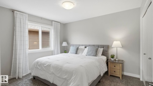 Serene primary bedroom featuring soft gray walls, abundant natural light from dual casement windows with sheer drapery, wood-finish nightstands with integrated task lighting, and plush bedding anchored by an upholstered headboard - 63 Rosemount Boulevard, Beaumont, AB - Indoor Photo Showing Bedroom