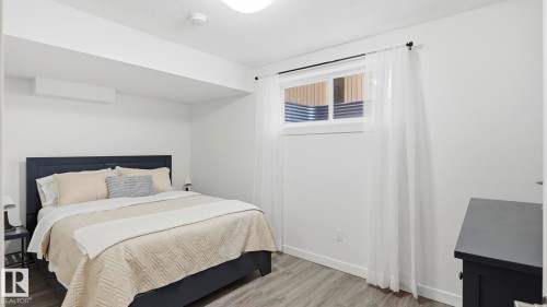 Bright secondary bedroom featuring a dark upholstered bed frame, neutral bedding, wood-finish flooring, and a large transom window with sheer curtains - 63 Rosemount Boulevard, Beaumont, AB - Indoor Photo Showing Bedroom