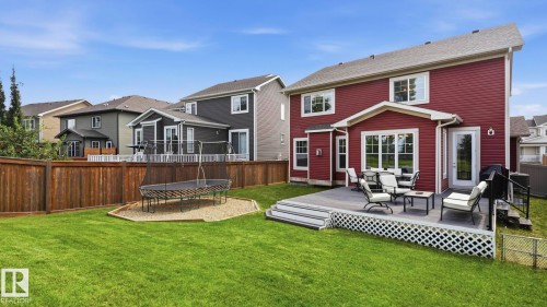 Striking red exterior with white trim and multiple windows overlooking a spacious backyard featuring a manicured lawn, composite deck with checkerboard railing, fire pit area with seating, and mature evergreen landscaping - 63 Rosemount Boulevard, Beaumont, AB - Outdoor With Deck Patio Veranda