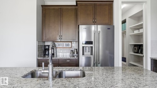 Stainless steel double-basin sink with a gooseneck faucet, light-toned stone-finish countertop, dark wood-finish cabinetry, stainless steel side-by-side refrigerator, and a recessed pantry with built-in shelving - 63 Rosemount Boulevard, Beaumont, AB - Indoor Photo Showing Kitchen With Double Sink With Upgraded Kitchen