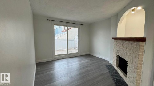Light-toned wood-finish flooring, white brick fireplace with dark mantel, dark stone hearth, arched wall niche with overhead lighting, and sliding glass door to an outdoor patio - 41 Lancaster Terrace Nw, Edmonton, AB - Indoor Photo Showing Other Room With Fireplace