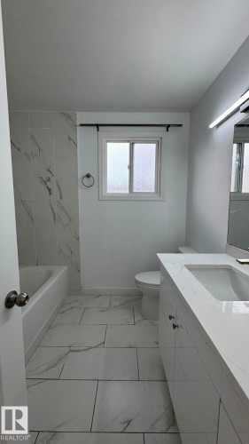 Bathroom featuring large format floor tiling, a white vanity with integrated dual sinks, a built-in bathtub, and a window with frosted glass - 41 Lancaster Terrace Nw, Edmonton, AB - Indoor Photo Showing Bathroom