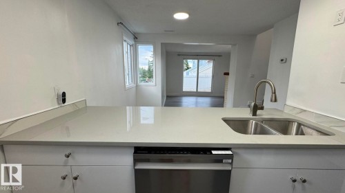 Light-toned solid surface countertop with an integrated double basin sink and gooseneck faucet, stainless steel dishwasher, and white cabinetry with brushed nickel hardware - 41 Lancaster Terrace Nw, Edmonton, AB - Indoor Photo Showing Kitchen With Double Sink