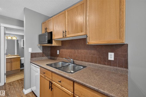 Wood-finish cabinetry with black hardware, stainless steel double basin sink, integrated white dishwasher, and brown tile backsplash - #4 4616 47 St, Leduc, AB - Indoor Photo Showing Kitchen With Double Sink