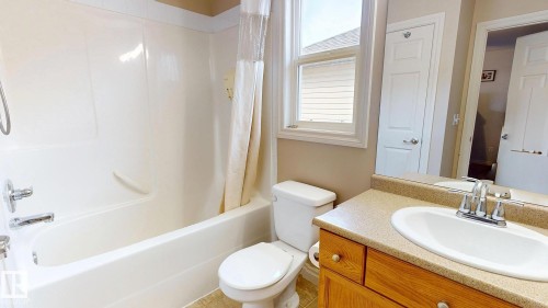 White bathtub with integrated shower, single-pane window, wood-finish vanity with a single sink, and mirror - 107 Hartwick Loop, Spruce Grove, AB - Indoor Photo Showing Bathroom