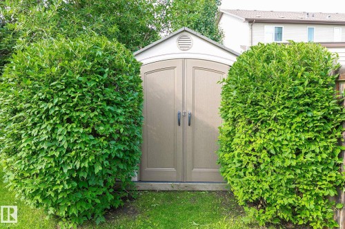 Versatile storage structure featuring a taupe double-door entry with arched roofline and ventilation detail, flanked by manicured emerald green hedges and set within a lush, level lawn landscape with mature tree canopy - 107 Hartwick Loop, Spruce Grove, AB - Outdoor