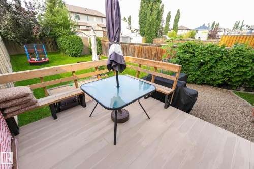Expansive wood-finish deck with built-in wooden bench seating, leading to a gravel path and fenced backyard with lush greenery and mature trees - 107 Hartwick Loop, Spruce Grove, AB - Outdoor With Deck Patio Veranda