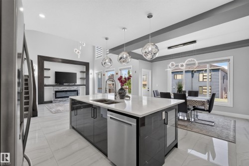 Open-concept living area featuring a large kitchen island with a white countertop and dark cabinetry, a built-in electric fireplace with stone detailing, recessed shelving, and large windows - 4703 41 Street, Beaumont, AB - Indoor Photo Showing Kitchen With Double Sink With Upgraded Kitchen
