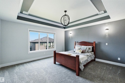 Tray ceiling with layered crown molding, contemporary chandelier, wall sconces, large window, and light grey carpeting - 4703 41 Street, Beaumont, AB - Indoor Photo Showing Bedroom