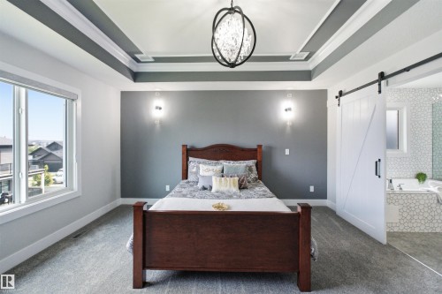 Spacious room featuring a tray ceiling with a contemporary chandelier, large picture window, and a sliding barn door entryway to an adjacent bathroom with a tiled soaking tub - 4703 41 Street, Beaumont, AB - Indoor Photo Showing Bedroom