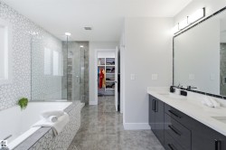 Modern bathroom featuring a dual vanity with white countertops and dark cabinetry, a large frameless mirror with integrated lighting, a spacious shower with glass enclosure, a built-in tub with mosaic tile surround, and polished floor tiling - 