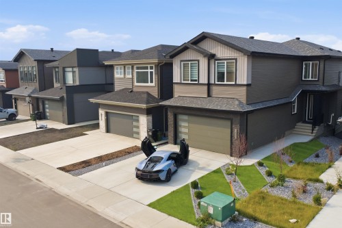 Modern two-story home featuring a sleek dark exterior with mixed siding finishes, double attached garage with contemporary paneling, and a professionally landscaped front yard with manicured lawn, decorative stone accents, and ornamental plantings - 5493 Kootook Road Sw, Edmonton, AB - Outdoor With Facade