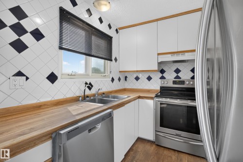 White cabinetry with wood trim, stainless steel appliances, wood-finish countertops, dual basin stainless steel sink, and a tiled backsplash - 2011 151 Avenue Nw, Edmonton, AB - Indoor Photo Showing Kitchen With Double Sink