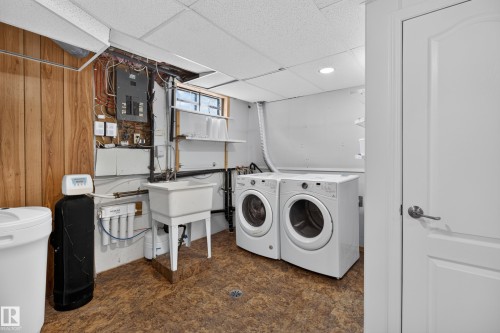 Dedicated laundry room featuring side-by-side front-load washers and dryers, cork-finish flooring, wood-paneled cabinetry with open shelving, utility sink with faucet, and water heater with mechanical systems - 2011 151 Avenue Nw, Edmonton, AB - Indoor Photo Showing Laundry Room