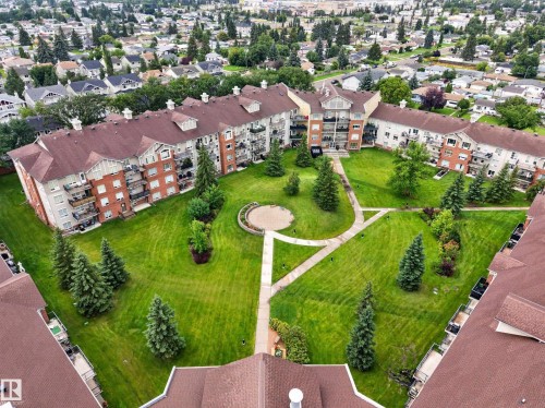 Brick courtyard community featuring expansive manicured grounds with mature evergreen landscaping, circular hardscaped pathways, and multi-unit residential architecture with pitched roof design - #205 6315 135 Av Nw, Edmonton, AB - Outdoor With View