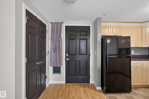 Wood-finish flooring, dark paneled entry door, kitchen with light wood cabinetry, dark countertops, and a black refrigerator - #205 6315 135 Av Nw, Edmonton, AB - Indoor Photo Showing Kitchen