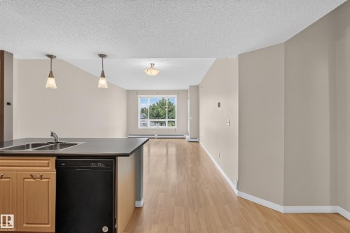 Galley kitchen featuring wood cabinetry, black appliances including a dishwasher, black countertops, pendant lighting, and wood-finish flooring throughout - #205 6315 135 Av Nw, Edmonton, AB - Indoor Photo Showing Kitchen With Double Sink