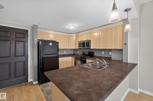 Contemporary kitchen featuring light wood cabinetry, stainless steel appliances including a black refrigerator and range, pendant lighting, and a spacious island with a dark quartz countertop and undermount sink - #205 6315 135 Av Nw, Edmonton, AB - Indoor Photo Showing Kitchen With Double Sink