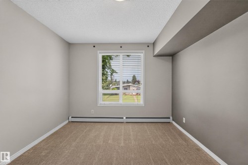 Bright, light-filled bedroom with expansive windows framing natural outdoor scenery, wood-finish flooring, neutral wall finishes, and contemporary ceiling details including a modern soffit element - #205 6315 135 Av Nw, Edmonton, AB - Indoor Photo Showing Other Room