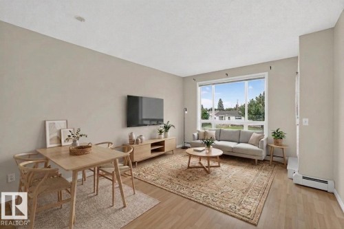 Open-concept living space featuring wood-finish flooring, a large picture window, and neutral wall tones - #205 6315 135 Av Nw, Edmonton, AB - Indoor Photo Showing Living Room