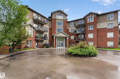 Multi-story building featuring brick and stucco exterior, multiple private balconies, a prominent main entrance with glass doors, and a circular landscaped island at the entrance - #205 6315 135 Av Nw, Edmonton, AB - Outdoor With Balcony With Facade