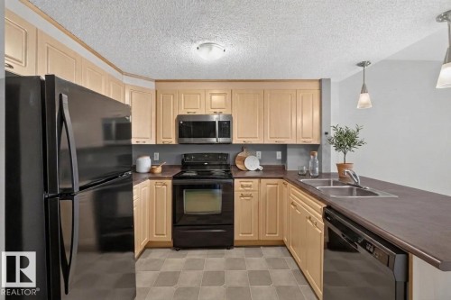 U-shaped kitchen featuring light-toned cabinetry, dark countertops, a double basin stainless steel sink, and black appliances - #205 6315 135 Av Nw, Edmonton, AB - Indoor Photo Showing Kitchen With Double Sink