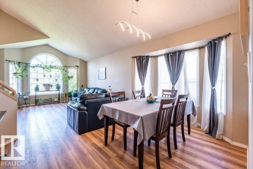 Vaulted ceiling over a living area with a large arched window, wood-finish flooring, a dining area with multiple windows, and a contemporary linear light fixture - 4705 43 Street, Beaumont, AB 