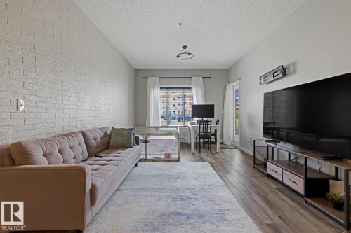 Textured accent wall, wood-finish flooring, recessed lighting, and a window with white drapes - #147 1818 Rutherford Rd Sw, Edmonton, AB - Indoor Photo Showing Living Room