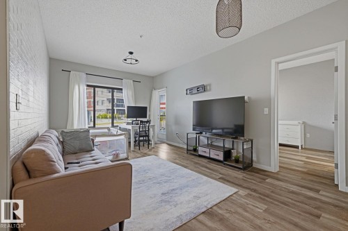 Wood-finish flooring throughout, complemented by a white brick accent wall - #147 1818 Rutherford Rd Sw, Edmonton, AB - Indoor Photo Showing Living Room