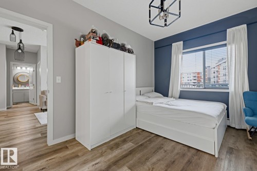 Wood-finish flooring throughout, ceiling-mounted light fixture, window with horizontal blinds, and a built-in vanity with an oval mirror in the adjacent space - #147 1818 Rutherford Rd Sw, Edmonton, AB - Indoor Photo Showing Bedroom