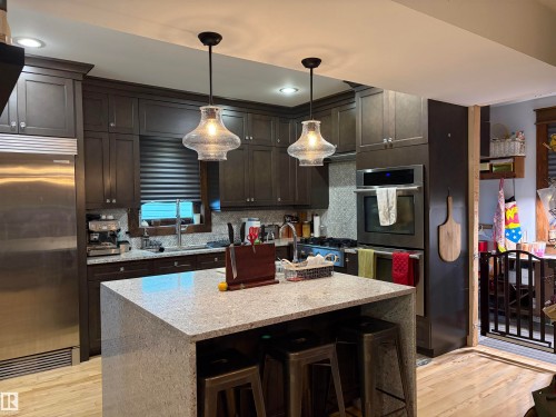 Kitchen featuring dark wood-finish cabinetry, stainless steel appliances, a center island with a light-toned countertop, and two pendant light fixtures - 10994 126 St Nw Nw, Edmonton, AB - Indoor Photo Showing Kitchen