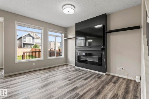 Wood-finish flooring throughout, modern electric fireplace with a black surround, two large windows, and a flush-mount ceiling light - 5210 23 Ave Sw, Edmonton, AB - Indoor Photo Showing Living Room With Fireplace