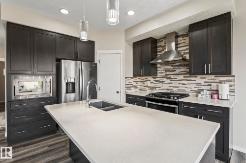 Kitchen featuring a central island with an undermount sink, dark cabinetry, stainless steel appliances, and a mosaic tile backsplash - 5210 23 Ave Sw, Edmonton, AB - Indoor Photo Showing Kitchen With Double Sink With Upgraded Kitchen
