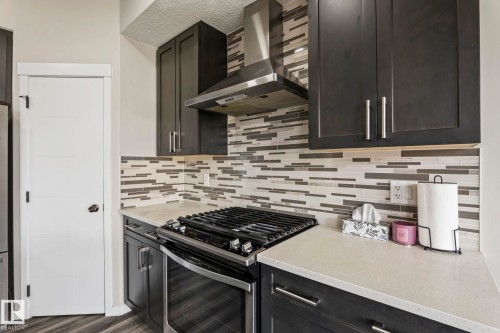 Stainless steel range with an overhead vent hood, dark cabinetry with bar pulls, mosaic tile backsplash, and light-toned countertops - 5210 23 Ave Sw, Edmonton, AB - Indoor Photo Showing Kitchen